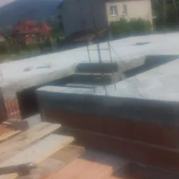 Świeżo wylany strop żelbetowy na murach z cegły, widoczne zbrojenia, w tle zabudowa jednorodzinna i zalesione wzgórza. Etap budowy domu.