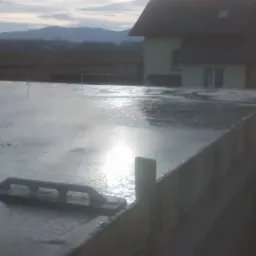 Świeżo wylany fundament budynku mieszkalnego z widoczną pacą do betonu, w tle dom i zalesione wzgórza pod pochmurnym niebem.