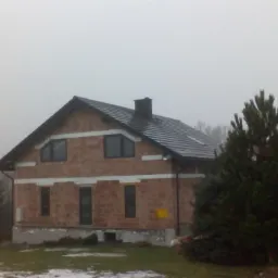 Surowy, dwukondygnacyjny budynek z czerwonej cegły z ciemnymi oknami i dachem z dachówki, otoczony zielenią, w pochmurny dzień. Widoczne fragmenty ocieplenia.