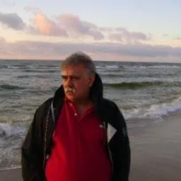 Mężczyzna w czerwonej koszuli i czarnej kurtce stoi na piaszczystej plaży na tle morza i zachodzącego słońca. Widoczne fale i pochmurne niebo.