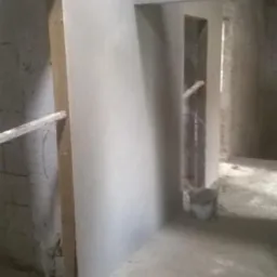 Wnętrze w trakcie prac wykończeniowych: świeżo położone, jasne tynki na ścianach, widoczne surowe otwory drzwiowe i okienne. W tle wiadro i surowy beton na podłodze.