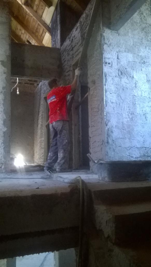 Tynkowanie maszynowe na poddaszu: mężczyzna w czerwonej koszulce wyrównuje świeży tynk na ścianie, widoczne drewniane elementy konstrukcyjne dachu.