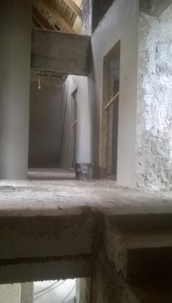 Wnętrze budynku w trakcie prac tynkarskich. Świeżo nałożony tynk na ścianach i kolumnach, surowe drewniane elementy konstrukcyjne widoczne w tle.