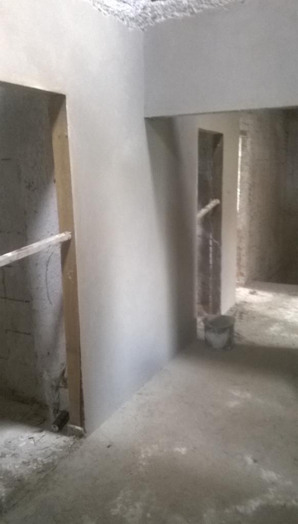 Wnętrze w trakcie prac wykończeniowych: świeżo położone, jasne tynki na ścianach, widoczne surowe otwory drzwiowe i okienne. W tle wiadro i surowy beton na podłodze.