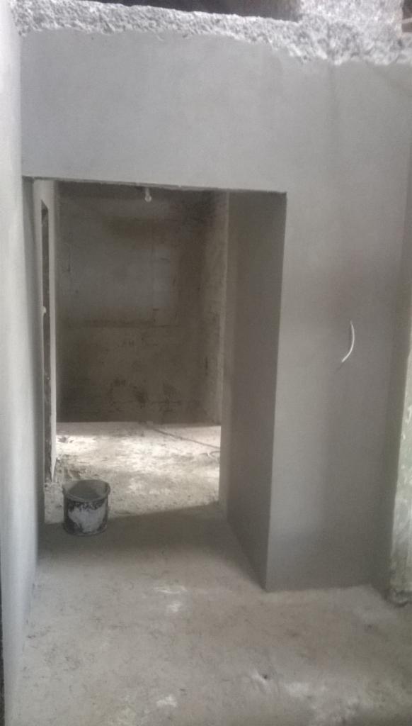 Wnętrze po tynkowaniu: gładkie, szare ściany, widoczne przejście do kolejnego pomieszczenia, wiadro na podłodze. Świeżo nałożone tynki maszynowe.