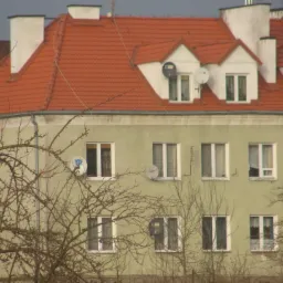 Wielorodzinny budynek z czerwoną dachówką, białymi kominami i antenami satelitarnymi na dachu. Widok zza drzew bez liści. Budynek otynkowany na oliwkowy kolor.