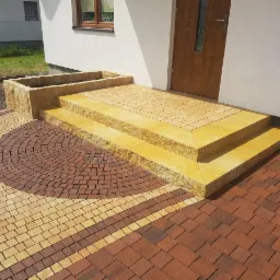 Wejście do domu z dwoma stopniami i donicą, wyłożone kostką w odcieniach brązu i beżu, ułożoną w geometryczne wzory.