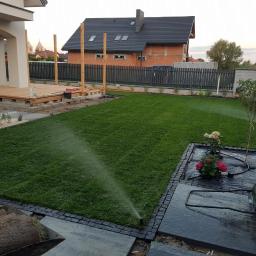 SM2 Mateusz Salamon - Nawadnianie świeżo rozłożonego trawnika z rolki, widoczne elementy systemu nawadniającego, w tle budynek w trakcie budowy i fragment drewnianego tarasu.