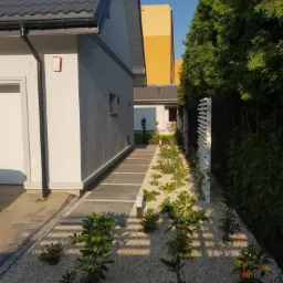 Nowoczesny ogród z białym żwirem, ścieżką z płyt betonowych i niskimi roślinami, oddzielony białym ażurowym płotem od wysokich tuj.