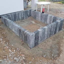 SM2 Mateusz Salamon - Niska obudowa rabaty kwiatowej wykonana z szarej kostki granitowej, z widocznymi śladami niedawnego montażu i ziemią wewnątrz, obok biały stolik z butelką Coca-Coli.