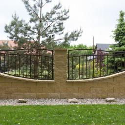 SM2 Mateusz Salamon - Ogrodzenie z bloczków betonowych w kolorze piaskowym z metalowymi przęsłami o łukowatym kształcie, widok na ogród i dom w tle.