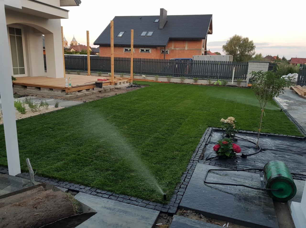 Nawadnianie świeżo rozłożonego trawnika z rolki, widoczne elementy systemu nawadniającego, w tle budynek w trakcie budowy i fragment drewnianego tarasu.
