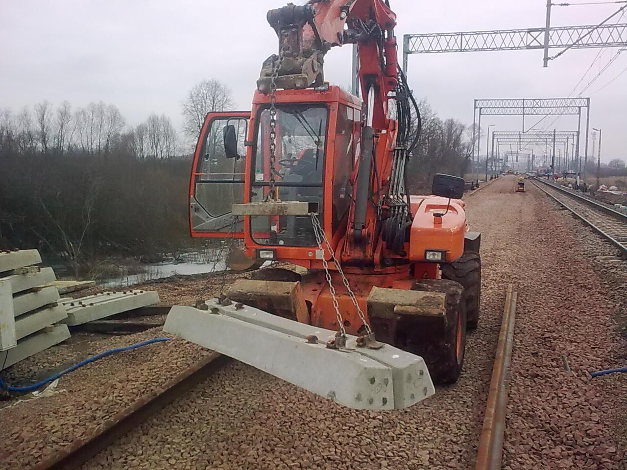 Pomarańczowa koparka kołowa z otwartymi drzwiami kabiny, podnosi betonowy podkład kolejowy na tle torów i sieci trakcyjnej. Prace na torach.