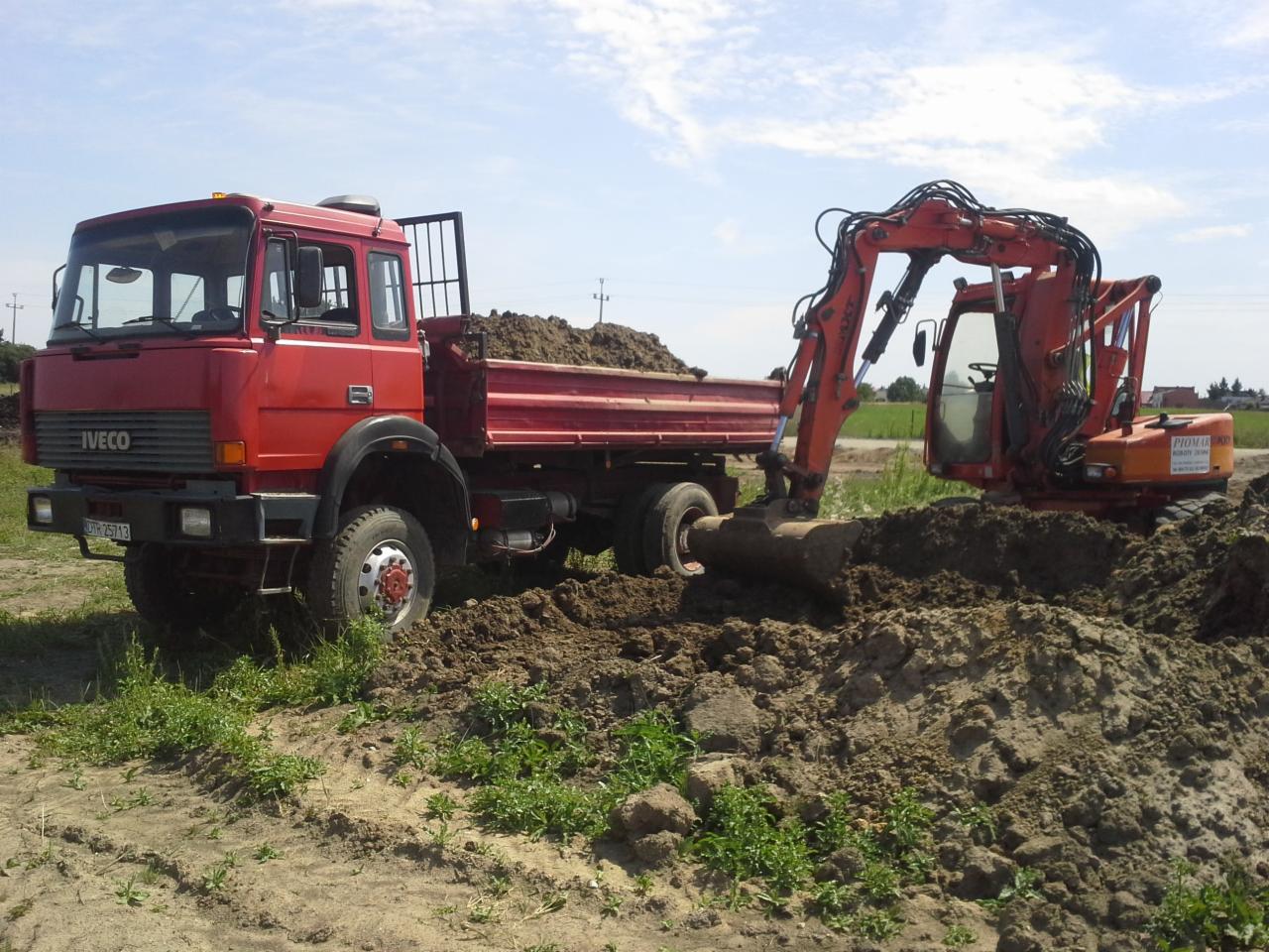 Czerwona ciężarówka IVECO i pomarańczowa koparka pracują na placu budowy, nabierając ziemię. W tle zielona łąka i błękitne niebo.