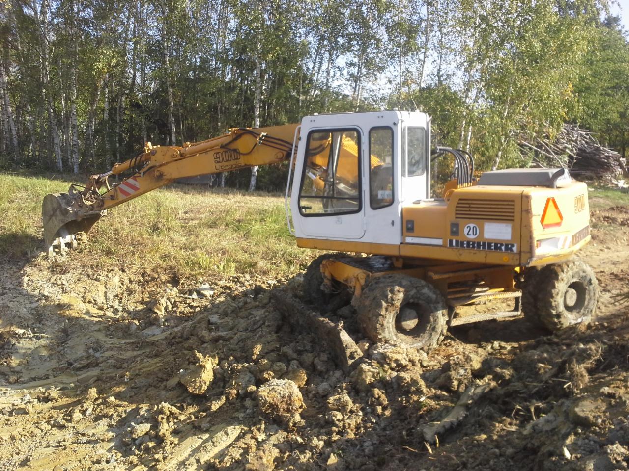 Żółta koparka Liebherr 900 na kołach, w trakcie prac ziemnych na terenie zalesionym, z widocznymi śladami opon w błocie i wykopanym dołem.