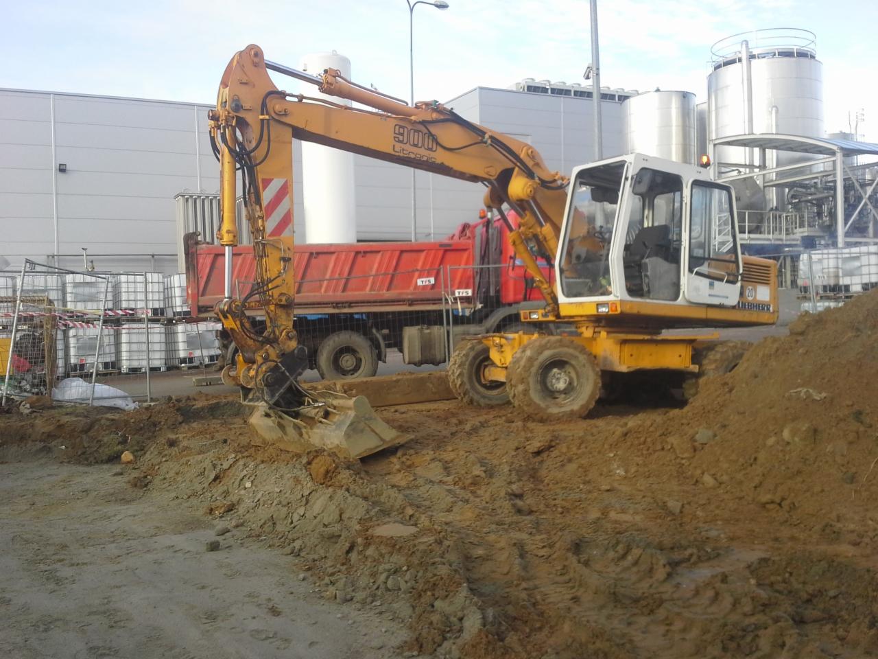 Żółta koparka kołowa Liebherr 900 przy wykopie, w tle czerwona wywrotka i białe zbiorniki przemysłowe.