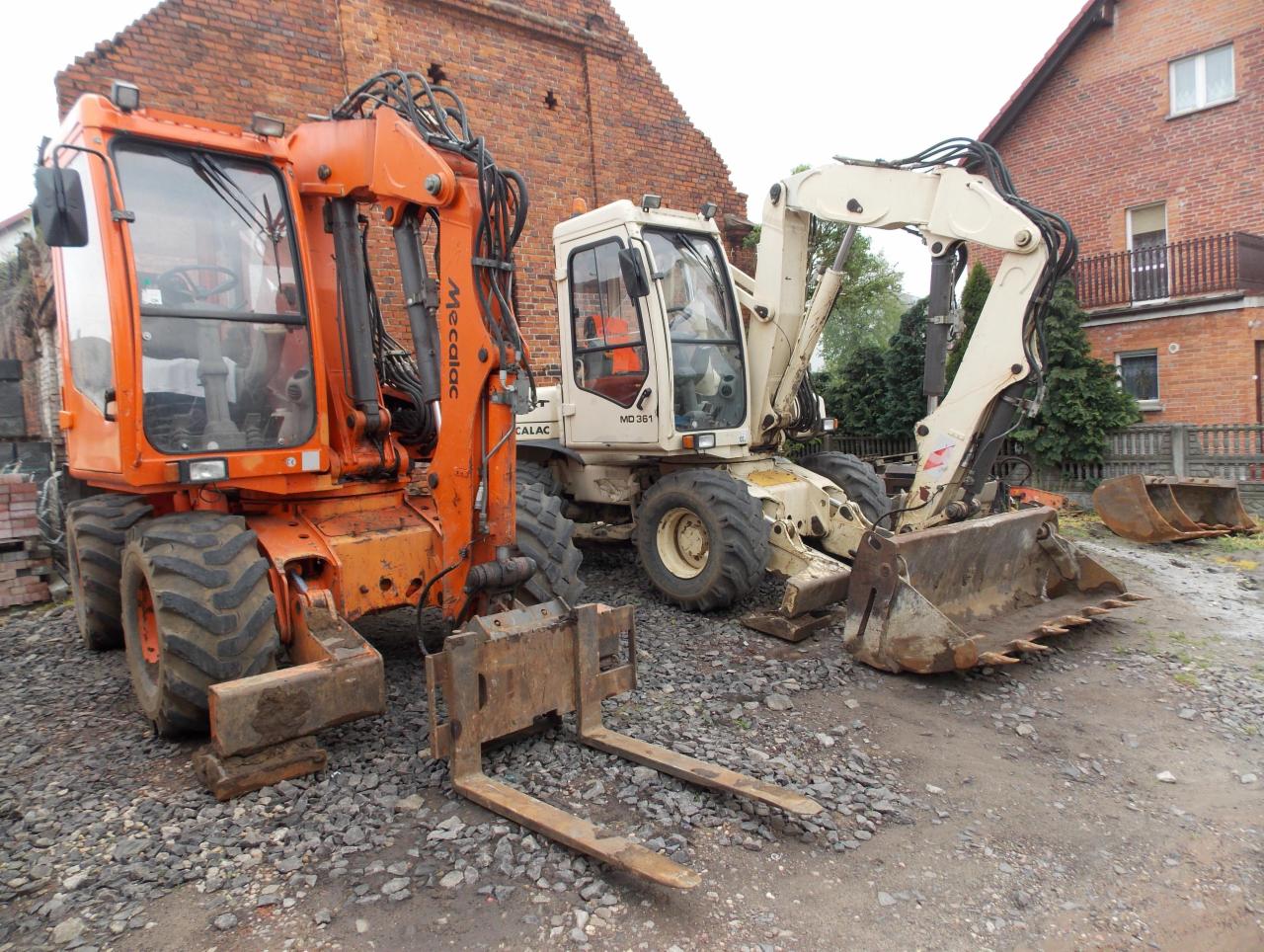 Dwa kołowe, używane, wielofunkcyjne koparki Mecalac: pomarańczowa z widłami i beżowa z łyżką, stoją na żwirowym podłożu przed budynkami z czerwonej cegły.