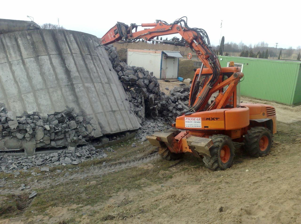 Pomarańczowy kombajn Piomar MXT kruszy betonowy silos. Gruz leży u podnóża konstrukcji. W tle kontenery i drzewa. Widoczne ślady opon na ziemi.