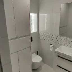 Nowoczesna, biała łazienka z podświetlaną wnęką, dekoracyjnym pasem płytek i minimalistyczną szafką pod umywalką z czarnymi uchwytami.