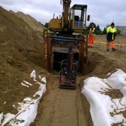 Firma Usługowo Handlowa Edward Krajcer - Żółta koparka umieszcza betonowy krąg w wykopie zabezpieczonym metalowymi szynami, ubijarka wibrująca na dnie wykopu, w tle dwóch robotników w odblaskowych ubraniach.