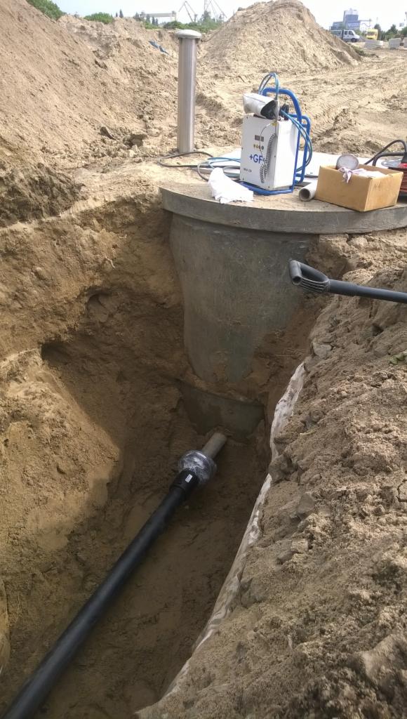 Wykonane wykopaliska, widoczny betonowy zbiornik, rura przyłączeniowa z uszczelnieniem, sprzęt spawalniczy +GF na górze zbiornika i zasypana ziemia w tle.
