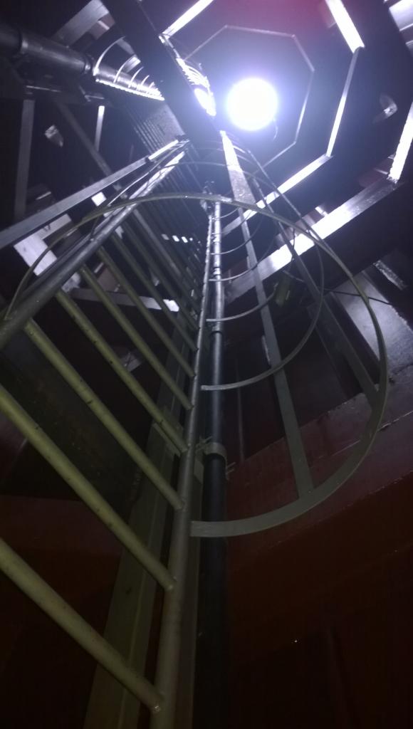 Wewnętrzna stalowa drabina z osłoną, prowadząca do oświetlonego otworu w betonowej konstrukcji. Widoczne pionowe rury i fragmenty drewnianej konstrukcji. Ujęcie od dołu.