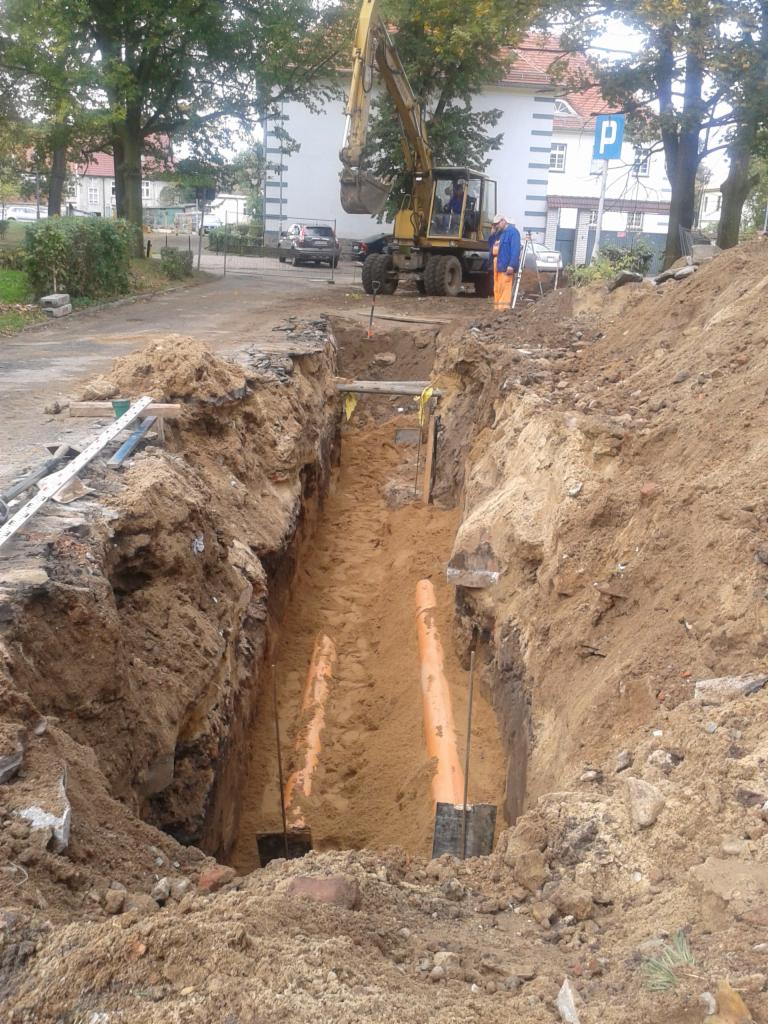 Wykop z pomarańczowymi rurami kanalizacyjnymi w ziemi, widoczny fragment koparki i pracownik w pomarańczowym kombinezonie, w tle budynek z znakiem drogowym P.