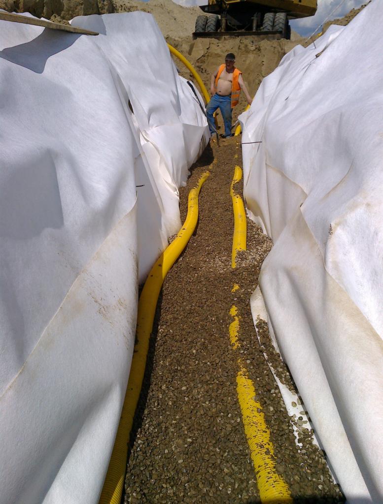 Wykop wyłożony białą geowłókniną, wewnątrz żółte rury drenarskie zasypane żwirem, na górze widoczny pracownik w kamizelce i koparka.