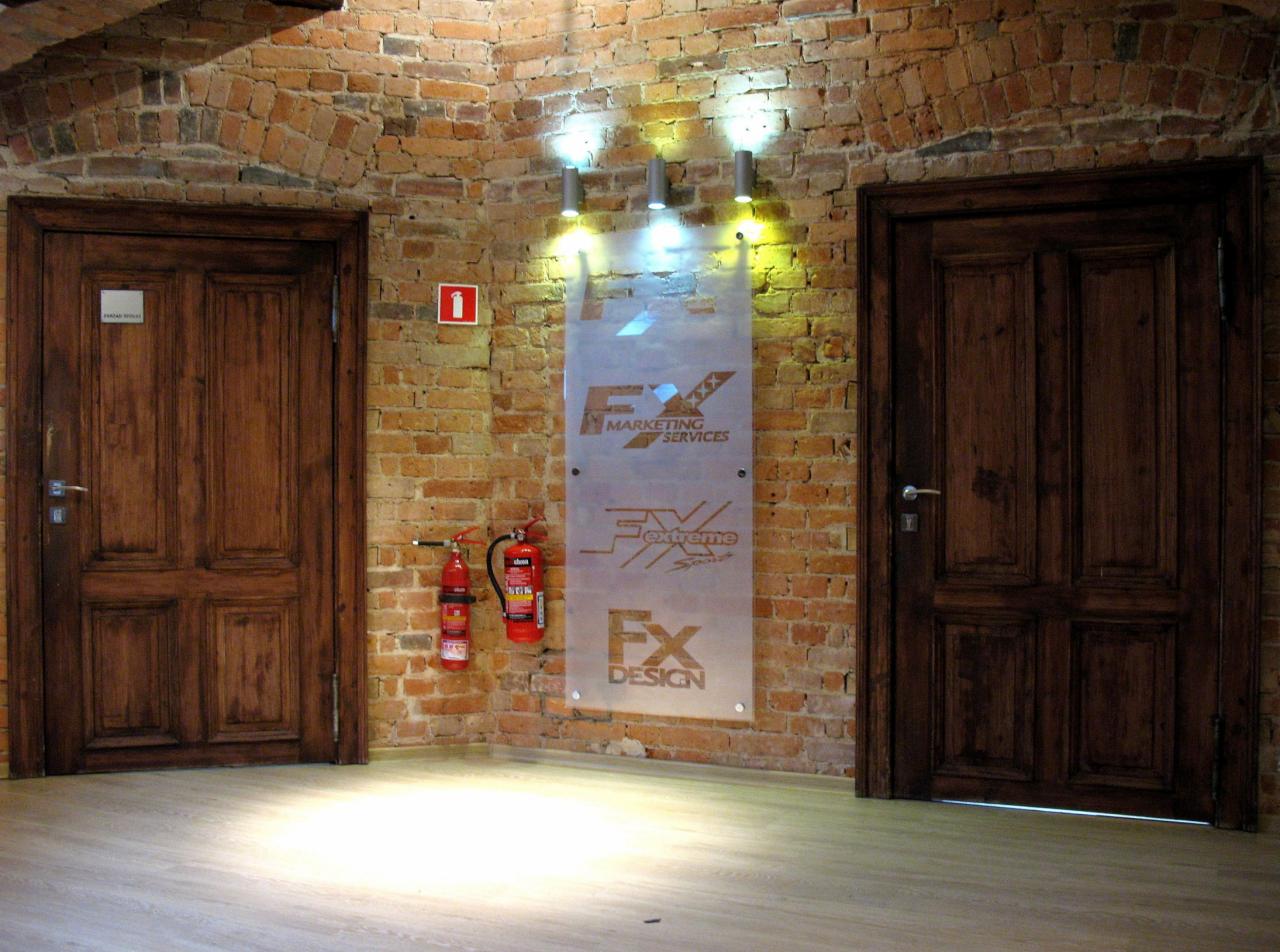 Wnętrze biura z dwiema drewnianymi drzwiami, ceglaną ścianą, logo firmy FX Marketing Services, gaśnicami i trzema lampami sufitowymi.