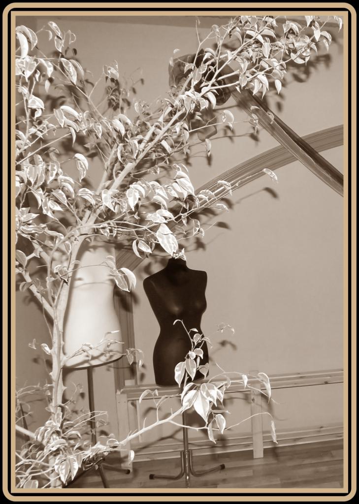 Sepia fotografia z dwoma manekinami krawieckimi, jeden biały, drugi czarny, otoczone roślinnością, tworzące artystyczną kompozycję w atelier.