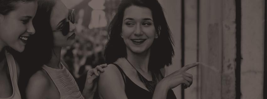 Trzy kobiety w odcieniach szarości, jedna z ciemnymi włosami wskazuje palcem, ubrana w sukienkę na ramiączkach i naszyjnik. Pozostałe kobiety ubrane w letnie bluzki.