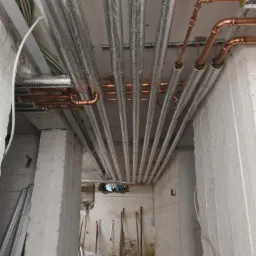 Szereg miedzianych rur instalacji grzewczej pod stropem, częściowo zaizolowanych srebrną folią, widoczne elementy mocujące, surowe betonowe ściany w tle.