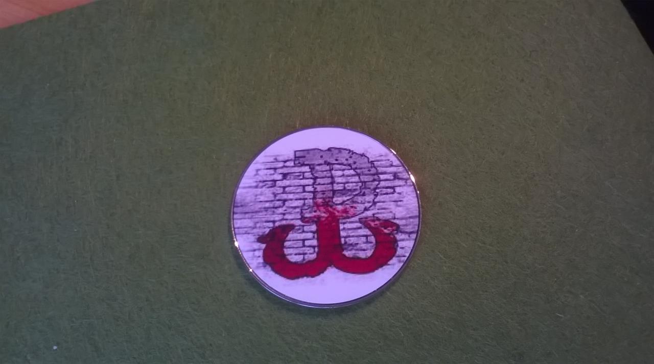 Okrągły magnes z symbolem Polski Walczącej na tle ściany z cegieł, leżący na zielonym tle.