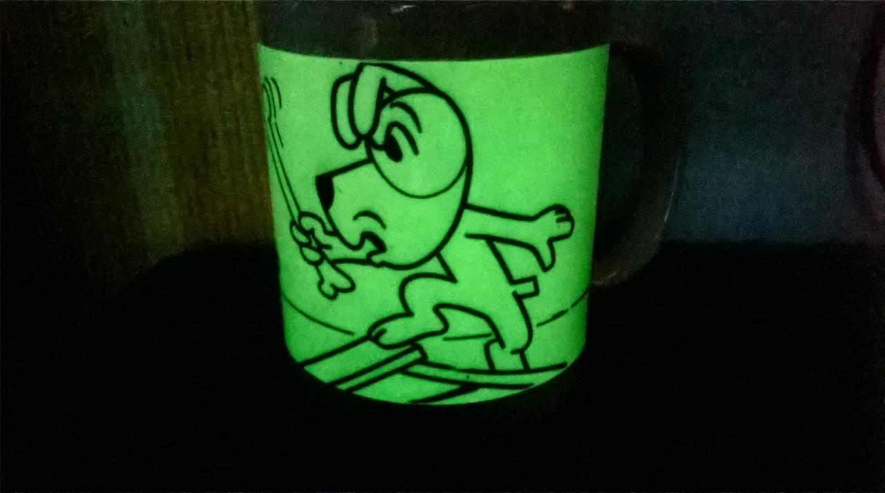 Świecący w ciemności kubek z czarnym konturem kreskówkowego psa trzymającego kość, ustawiony na ciemnym tle.