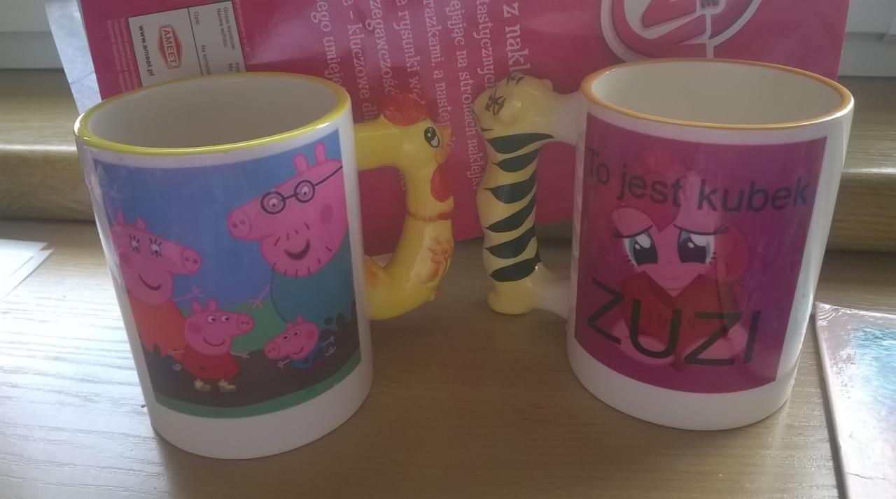 Dwa białe kubki z nadrukiem: jeden z bohaterami bajki 'Świnka Peppa', drugi z 'My Little Pony', oba z żółtymi obwódkami i uchwytami w kształcie zwierząt.