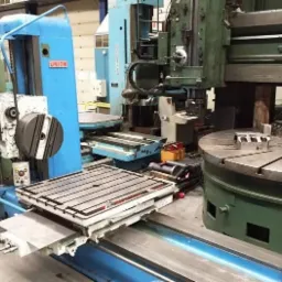 Rozległa hala z dwoma dużymi, stacjonarnymi obrabiarkami do metalu, jedna w kolorze niebieskim z logo 'UNION', druga w odcieniach zieleni, z metalowymi elementami obrabianymi na okrągłym stole.