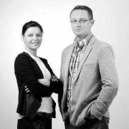 Portret biznesowy w czerni i bieli: uśmiechnięta kobieta z założonymi rękami i mężczyzna w marynarce, oboje patrzą w obiektyw na jasnym tle.