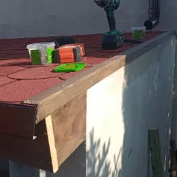 Montaż obróbki blacharskiej na drewnianej konstrukcji z użyciem wkrętarki, młotka i pojemników z wkrętami na tle czerwonego dachu z gontu bitumicznego.