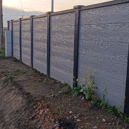 PRO-MONTAŻ - Szary, panelowy płot z elementami imitującymi drewno, w tle słup oświetleniowy i zachodzące słońce. Na pierwszym planie ziemia z roślinnością.