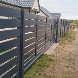 PRO-MONTAŻ - Nowoczesne ogrodzenie panelowe w Lipnie, z poziomymi szczeblami w kolorze grafitowym, zamontowane wzdłuż posesji. W tle widoczny dom i transport elementów ogrodzenia.