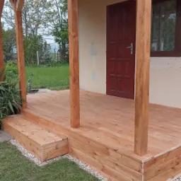 Drewniany taras z zadaszeniem przy budynku, wykończony drobnymi kamieniami, widok na brązowe drzwi wejściowe i okno.