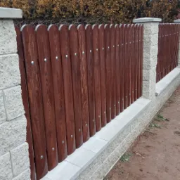 Nowoczesne ogrodzenie z drewnianych sztachet w kolorze ciemnego brązu, przymocowanych do betonowych słupk&oacute;w i podmur&oacute;wki, z widocznymi srebrnymi śrubami, na tle zielonych krzew&oacute;w i częściowo...