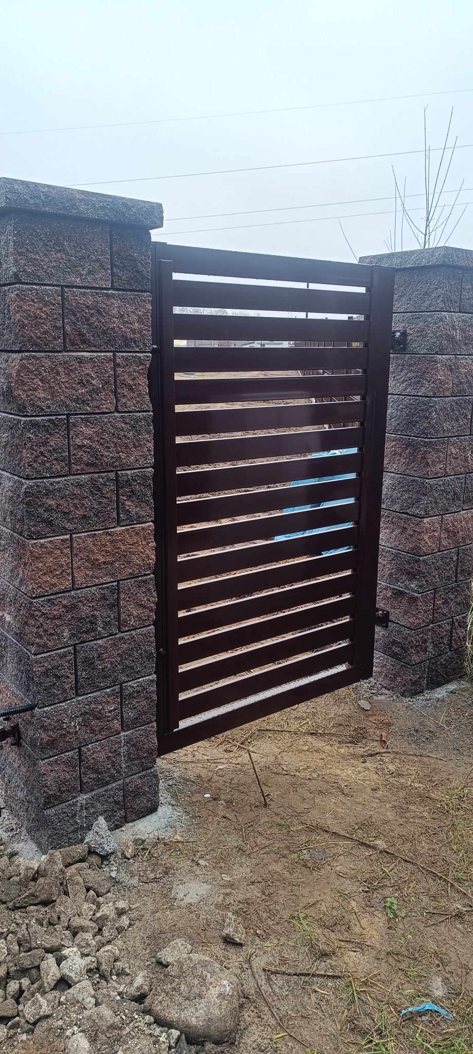 Nowoczesna brama metalowa z poziomymi szczeblami, osadzona między dwoma murowanymi słupkami z cegły klinkierowej. Widoczne niedokończone prace ziemne.