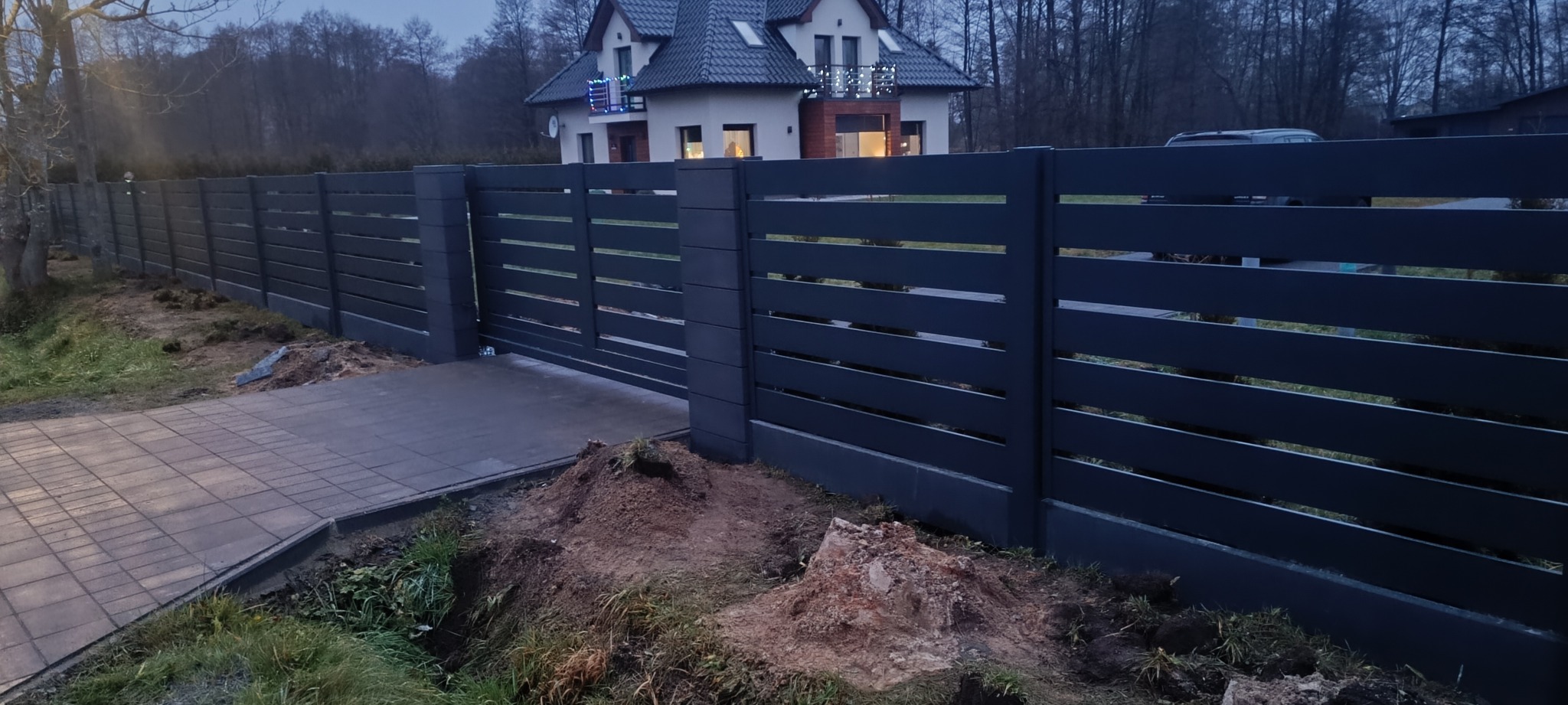 Nowoczesne, poziome przęsła grafitowego ogrodzenia z betonowymi słupkami na tle domu z podświetlonym balkonem i brukowanym podjazdem. Widok z poziomu gruntu.