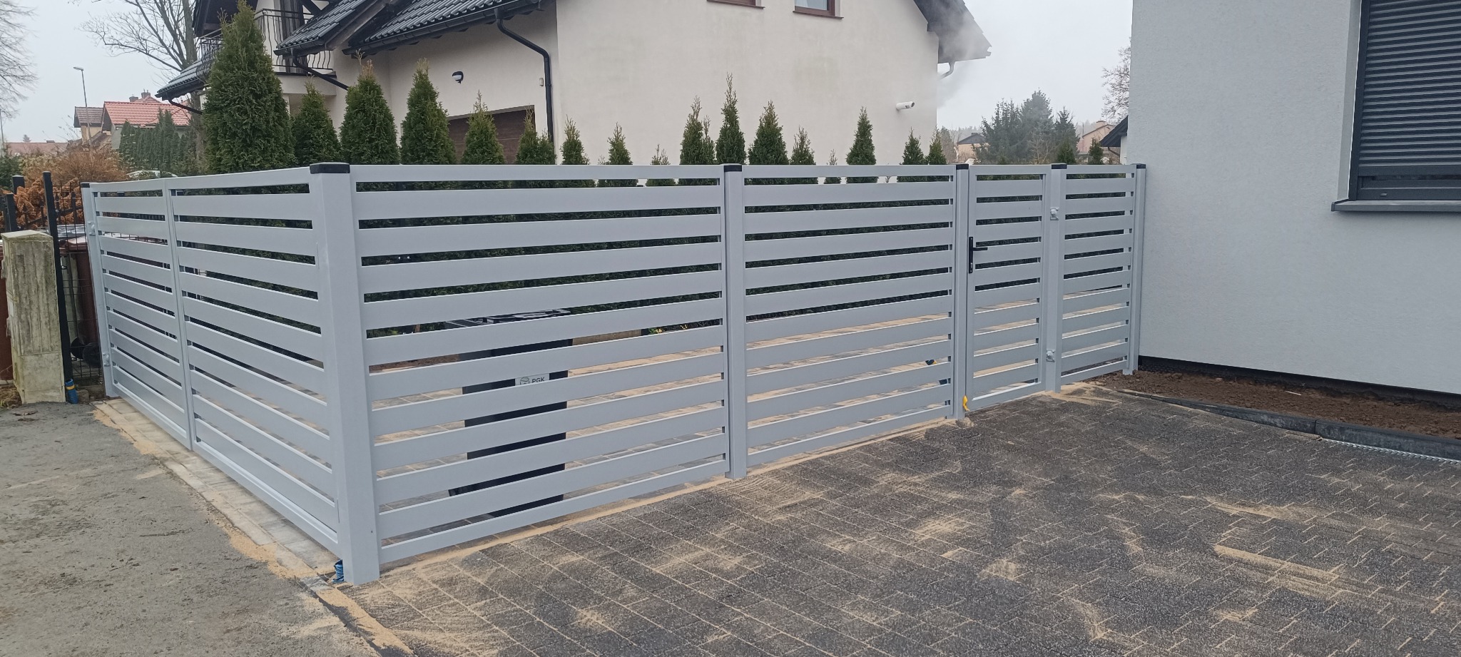 Nowoczesne, poziome, szare ogrodzenie panelowe z bramą wjazdową, widok z zewnątrz posesji. Tło stanowi dom z tujami.