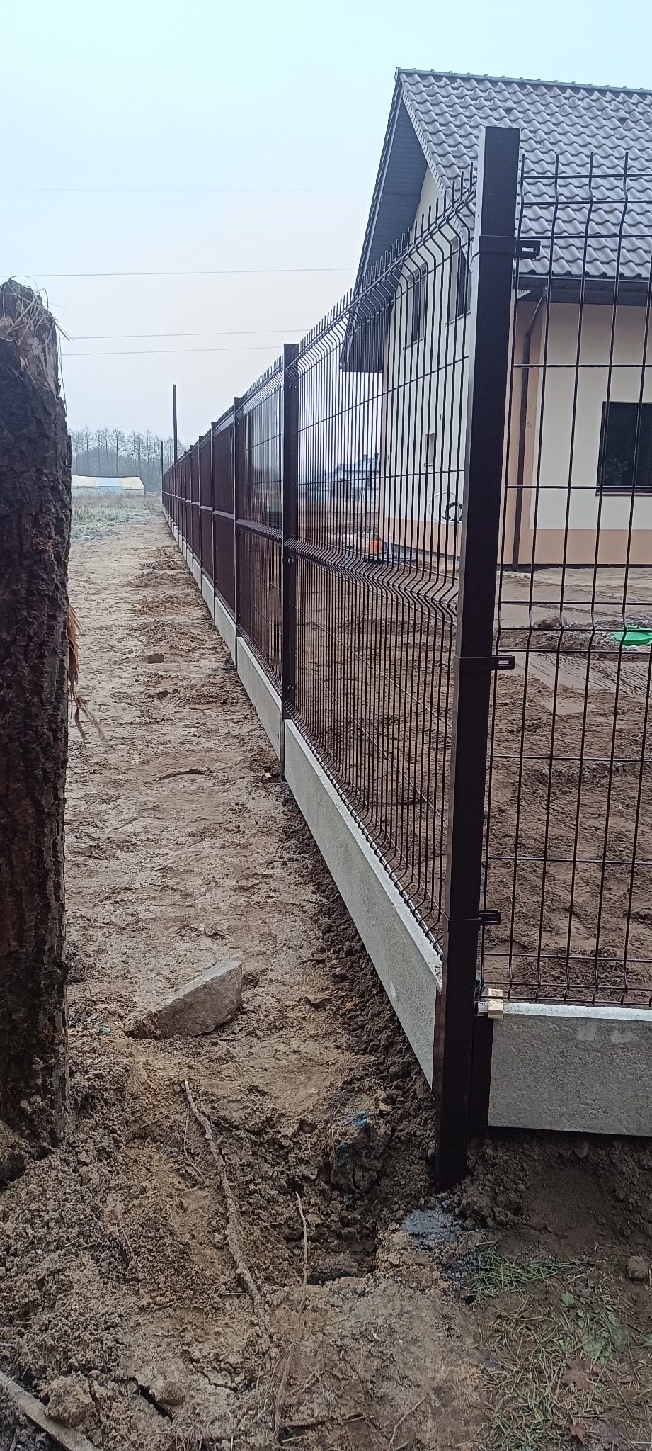 Długi, brązowy panel ogrodzeniowy z betonową podmurówką, ciągnący się wzdłuż posesji. Widok z boku, w tle dom z ciemnym dachem i pochmurne niebo.