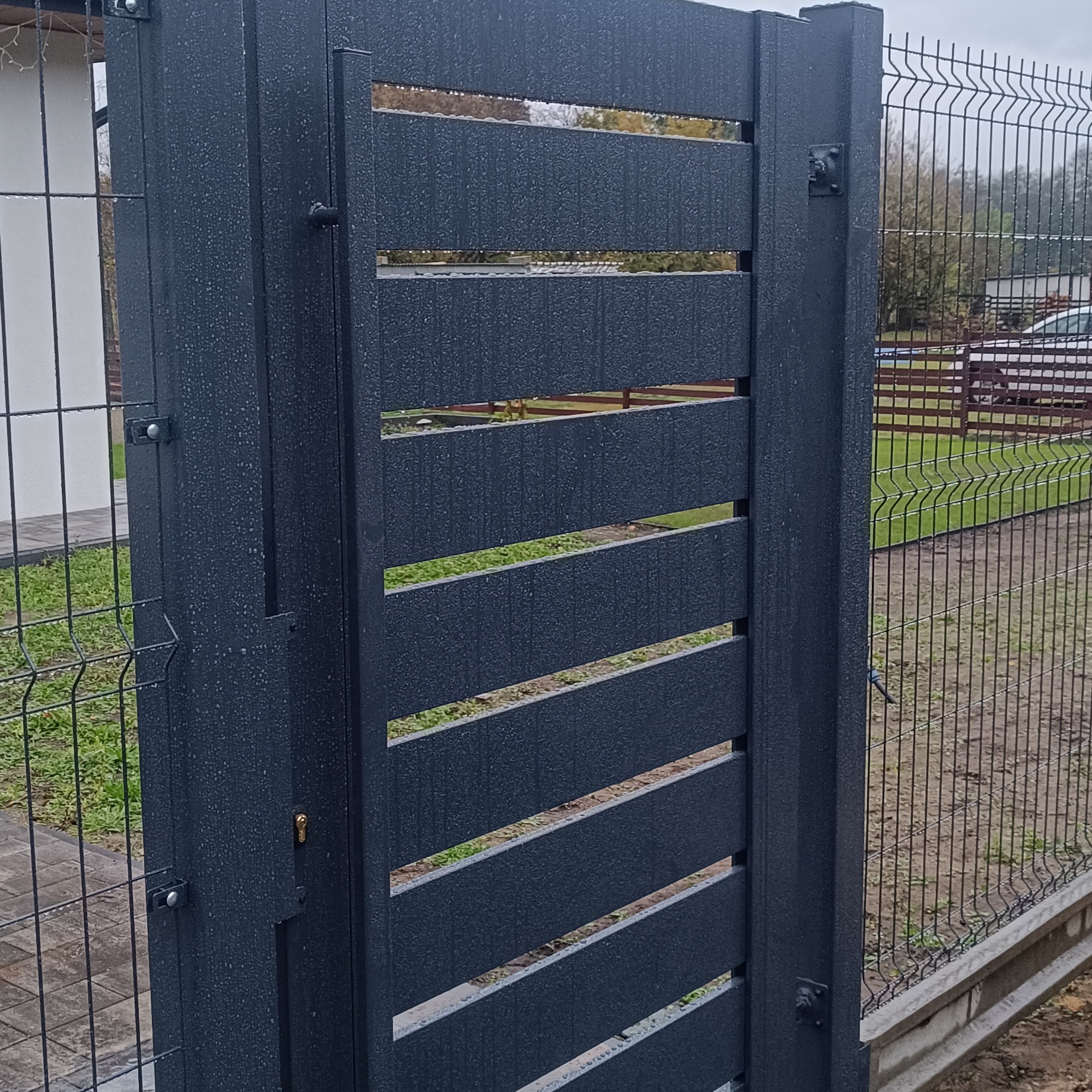 Ciemna, metalowa furtka z poziomymi szczebelkami, połączona z panelem ogrodzeniowym. Widoczne krople deszczu na powierzchni. Tło: dom i zieleń.