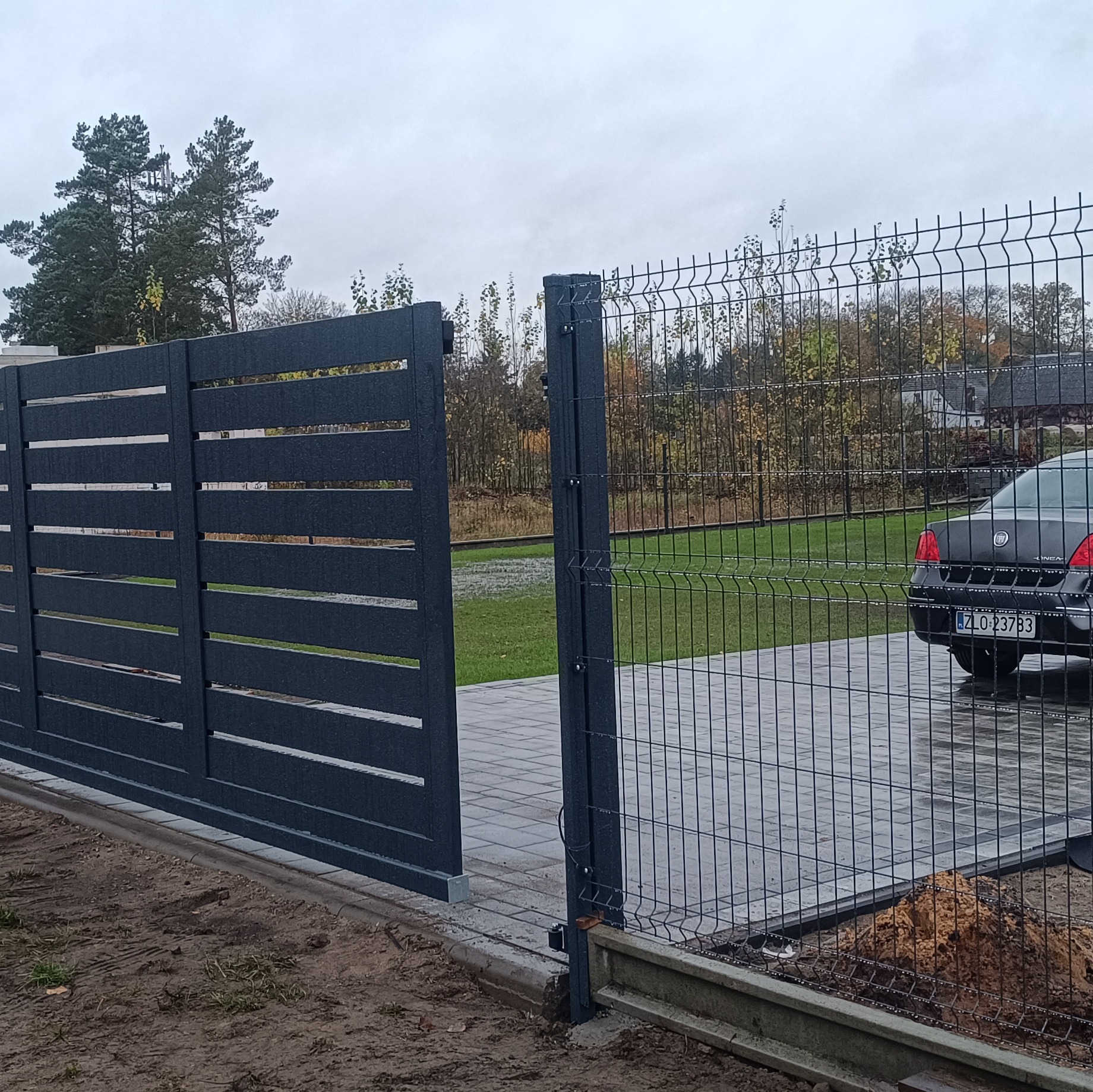Nowoczesna, grafitowa brama przesuwna z poziomymi panelami i fragmentem ogrodzenia panelowego. Widoczny samochód za ogrodzeniem. Pochmurny dzień.