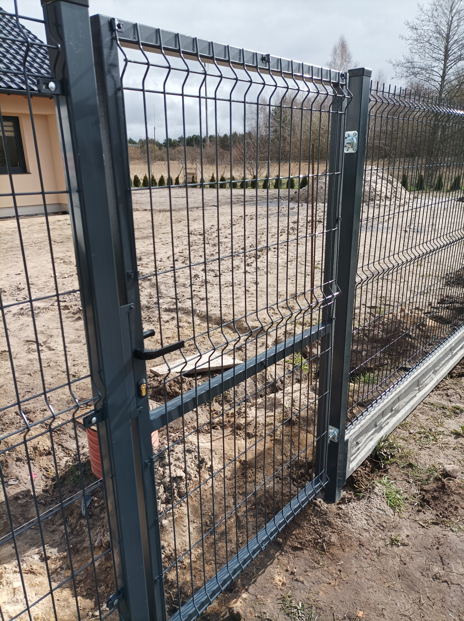 Fragment posesji z nowym, grafitowym ogrodzeniem panelowym z bramką wejściową, widoczne słupki i podmurówka betonowa, teren w trakcie prac ziemnych.