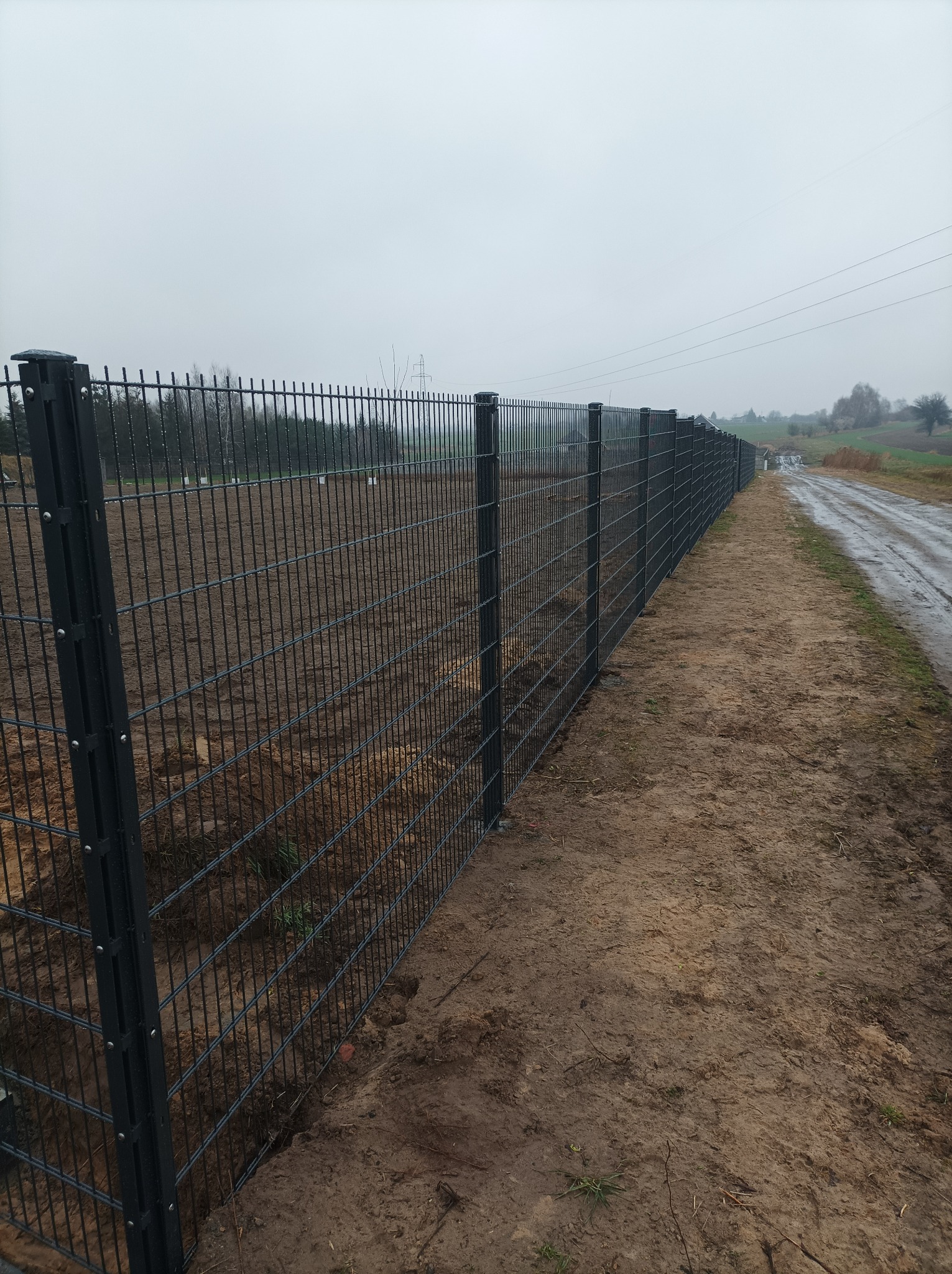 Długi, ciemnoszary płot z siatki panelowej, zamontowany wzdłuż błotnistej drogi polnej na tle pochmurnego nieba i drzew bez liści.